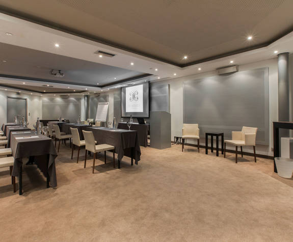 Spacious and modern Ismini meeting room at The Excelsior Hotel, arranged with long conference tables, cream upholstered chairs, projection screen, flip chart, water bottles, and a beverage station in a neutral setting