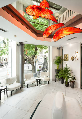 Bright, modern lobby area with large windows, stylish white seating, lush green plants, and striking orange sculptural ceiling lights, offering a welcoming view to the lively street outside