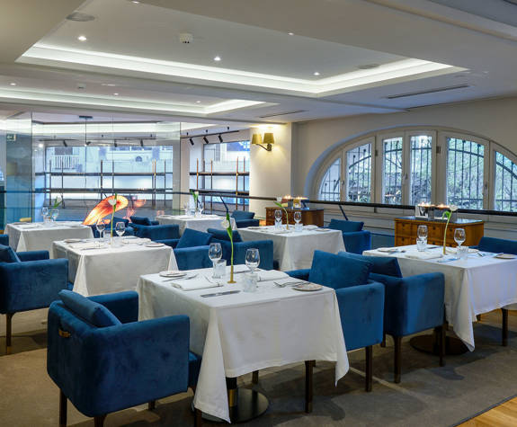 Elegant Mezzanine setup at The Excelsior Hotel arranged for business meetings, featuring square tables with crisp white linens, blue velvet chairs, neatly set glassware, and a modern, light-filled atmosphere