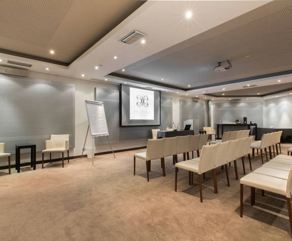Ismini meeting room at The Excelsior Hotel configured for theater-style seating with rows of cream upholstered chairs facing a projection screen, flip chart, and beverage station in a modern neutral-toned space