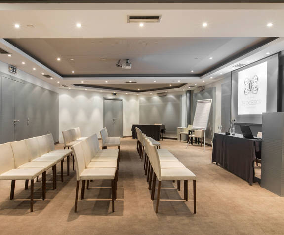 Ismini meeting room at The Excelsior Hotel, arranged in theater style with rows of cream chairs facing a projection screen and presenter’s table in a spacious, modern, and well-lit conference setting