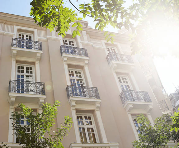 Sun-dappled façade of The Excelsior Hotel, with ornate balconies and tall windows, framed by lush greenery and bright natural light for a fresh, welcoming atmosphere
