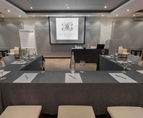 Excelsior Hotel’s Ismini meeting room featuring U-shaped table setup with notepads, pens, water bottles, wine glasses, projection screen, flip chart, and cream upholstered chairs in a modern, softly lit environment