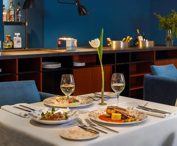 Elegant Mezzanine business meal setup at The Excelsior Hotel, featuring blue velvet chairs, crisp white tablecloths, gourmet dishes, glasses of white wine, and a single white tulip centerpiece