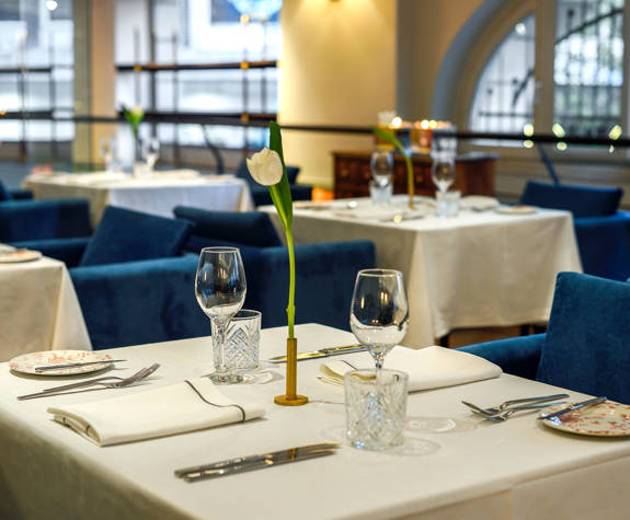 Sophisticated Mezzanine business dining at The Excelsior Hotel, with blue velvet chairs, crisp white table linens, crystal glassware, and single white tulip centerpieces on each table