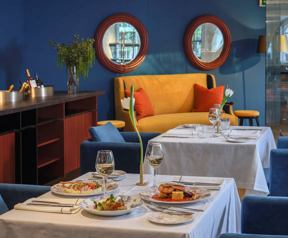 Refined Mezzanine dining at The Excelsior Hotel with blue velvet seating, white tablecloths, tulip centerpieces, white wine, and artfully plated gourmet dishes in a sophisticated blue and orange setting